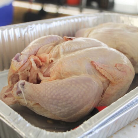 A raw chiken in a foil pan with the leggs trussed together.