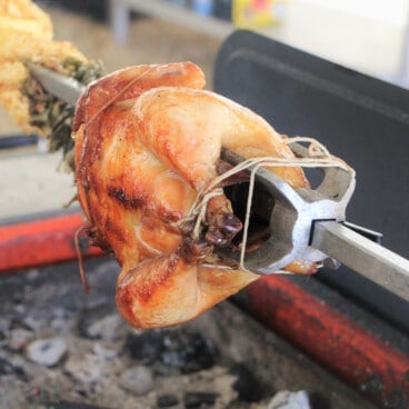 A rich golden brown cooked chicken is on a spit over charcoal.