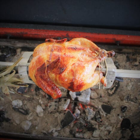 A golden brown whole cooked chicken on a spit over charcoal.