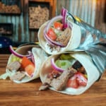 Three ready to eat lamb kebab wraps are stacked on a wooden bench.