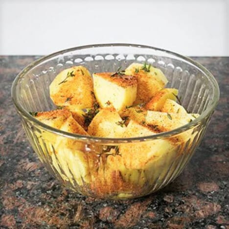 A glass bowl filled with cut potatoes and topped with a range of seasonings.