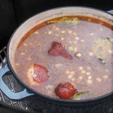 A Dutch oven with simmering beans and ham hocks on a smoker.