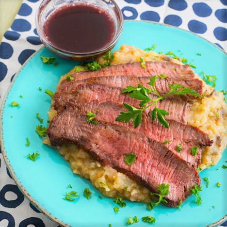Sliced flat iron steak sprinkled with parsley rests atop mashed potatoes on a blue plate, with a bowl of red wine sauce on the side.