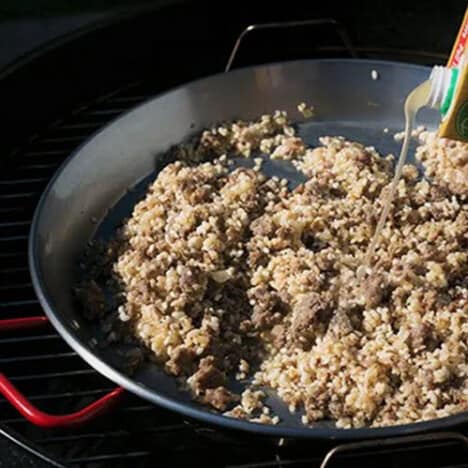 Ground sausage and rice in a paella pan with stock being poured in.
