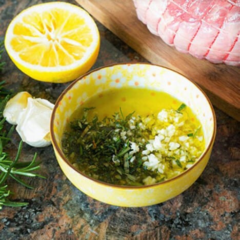 A small yellow bowl, sitting next to a halved lemon and a piece of raw lamb, holds oil, rosemary, and garlic.