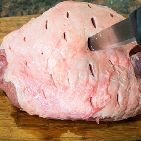 A raw leg of lamb is being pierced with a knife to create tiny slits over the surface.
