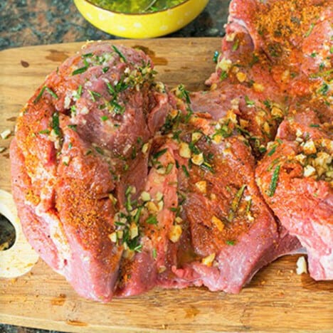 A butterflied leg of lamb, on a wooden cutting board, is covered with fresh herbs and red rub.