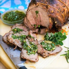 A smoked leg of lamb, drizzled with green chimichurri sauce, sits sliced on a wooden cutting board.