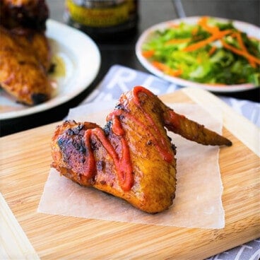 A single grilled chicken wing drizzled with hot sauce sits on a wooden cutting board with salad in the background.