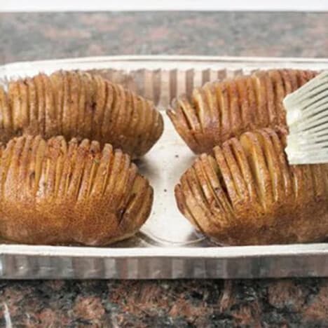 Four Hasselback prepared potatoes being brushed with melted butter.