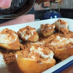 In a white baking dish, half pears with their stuffing and a biscuit crumble are having maple syrup poured over them.