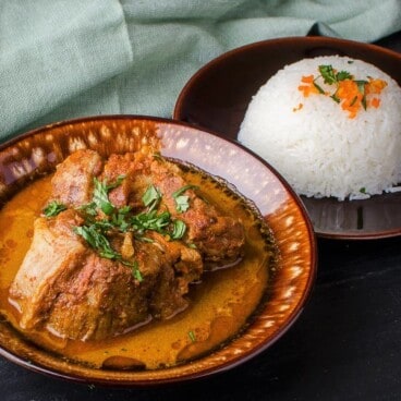 A serving of a large piece of pork in a Thai red curry gravy next to a side of steamed rice.