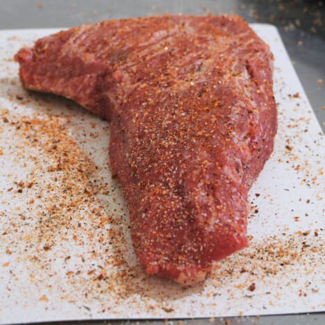 A raw tri-tip sitting on a white chopping board covered with a beef rub.