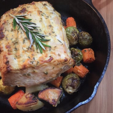 Looking down on a roast pork loin garnished with a sprig of rosemary and sitting onroast vegetables in a skillet.