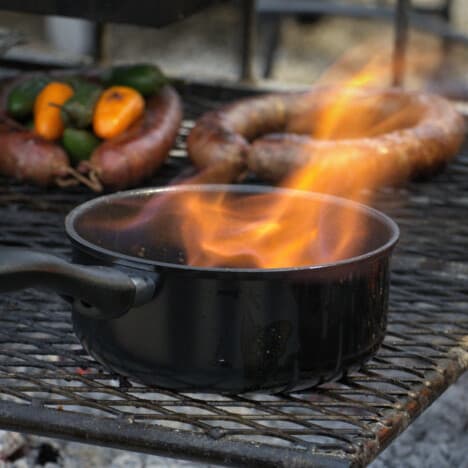 Saucepan with a flame coming out of it on a grill with sausages in the background.