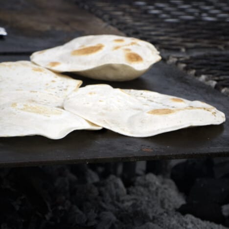 Tortillas on a flat top grill over charcoal being toasted to a golden brown.