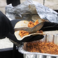 Black gloved hands scooping a spoonful of chorizo and egg mixture to fill a tortilla.