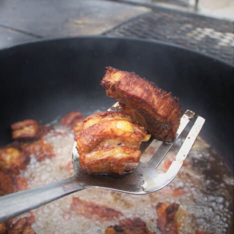 Two pieces of Chicharron being held on a fork in front of a Dutch oven with additional pieces cooking.